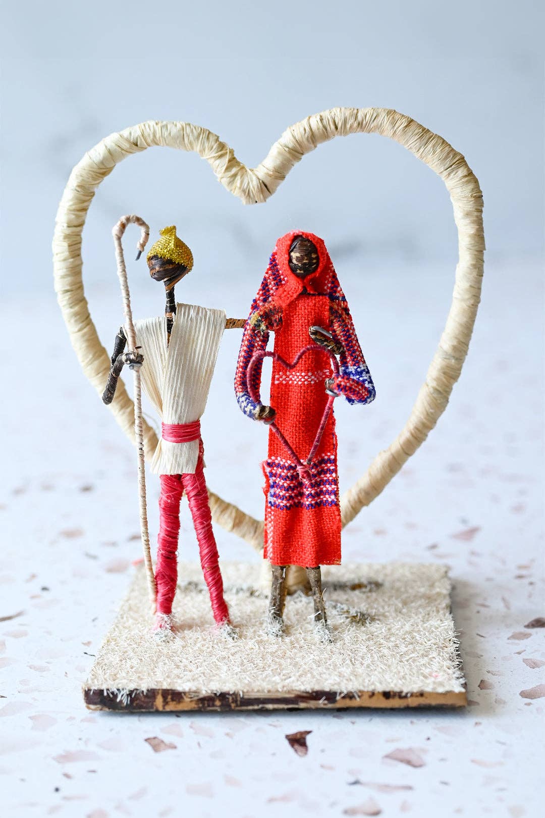 Two dolls in traditional attire standing under a heart-shaped frame on a textured surface.